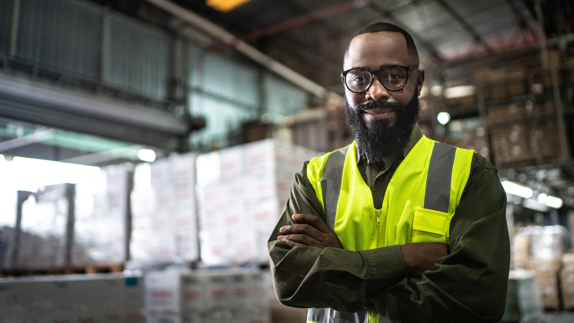 A smiling worker in a high visibility vest stands in front of palettes in a warehouse.