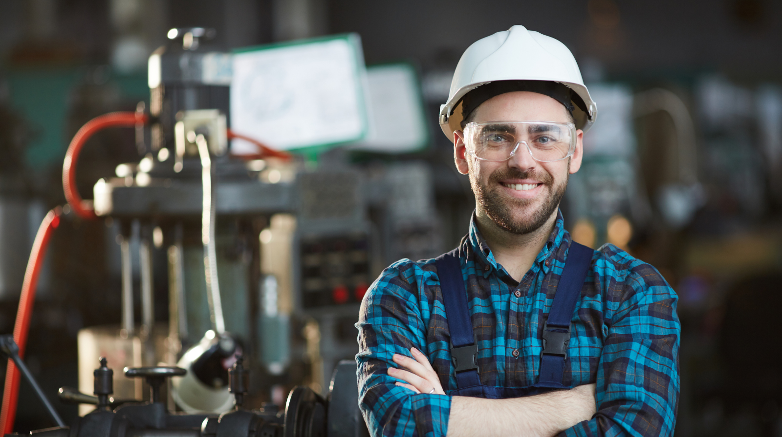 Smiling industrial worker wearing a white hard hat, clear safety glasses, and a blue plaid shirt with suspenders, standing with arms crossed in a manufacturing or industrial facility.