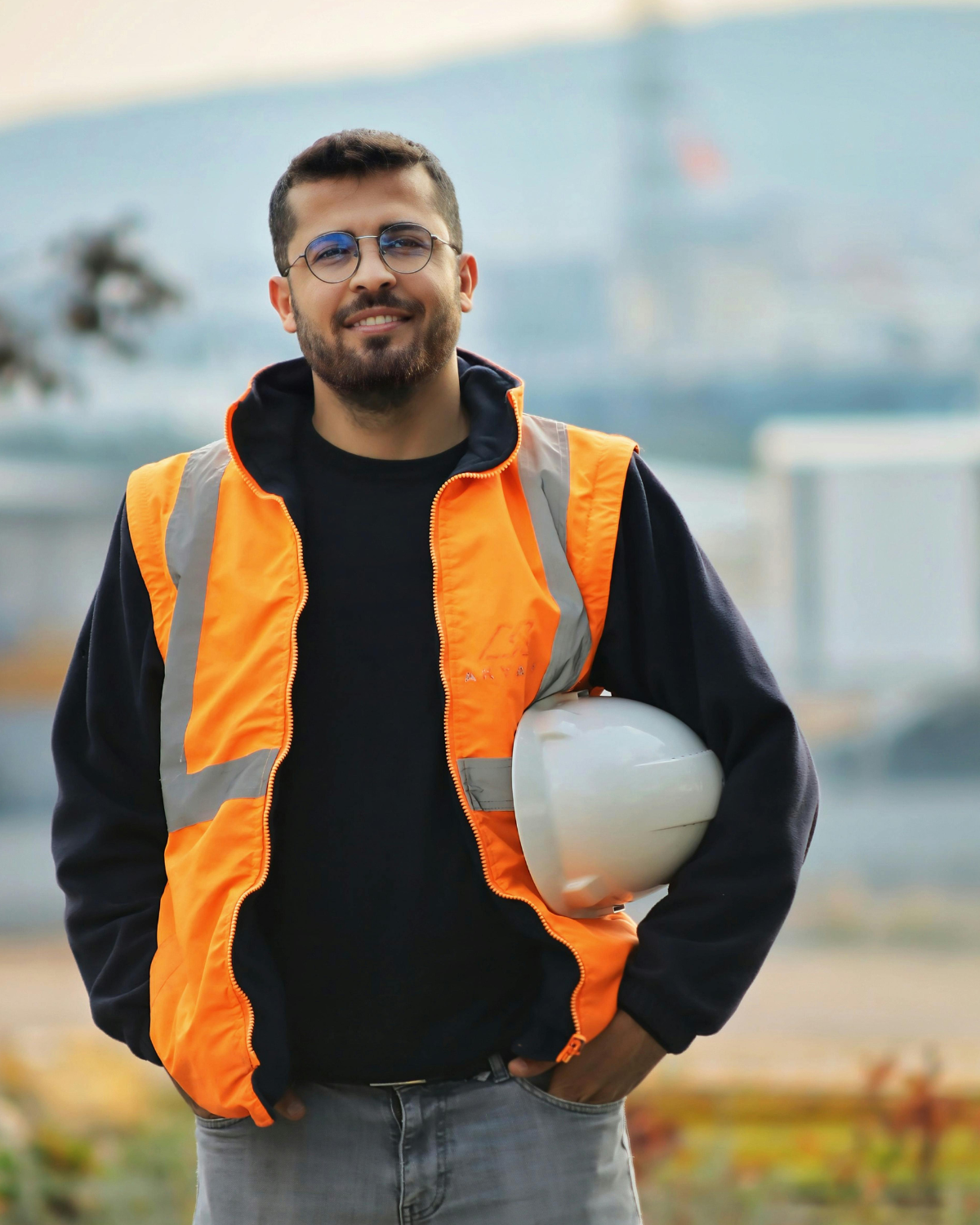 Construction worker wearing a high-visibility safety vest and glasses holds a hard hat while standing at a job site, smiling confidently.