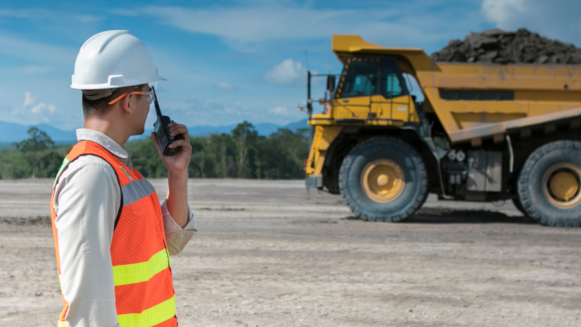 Construction worker wearing a hard hat and safety vest uses a radio while standing near a large dump truck at a job site.