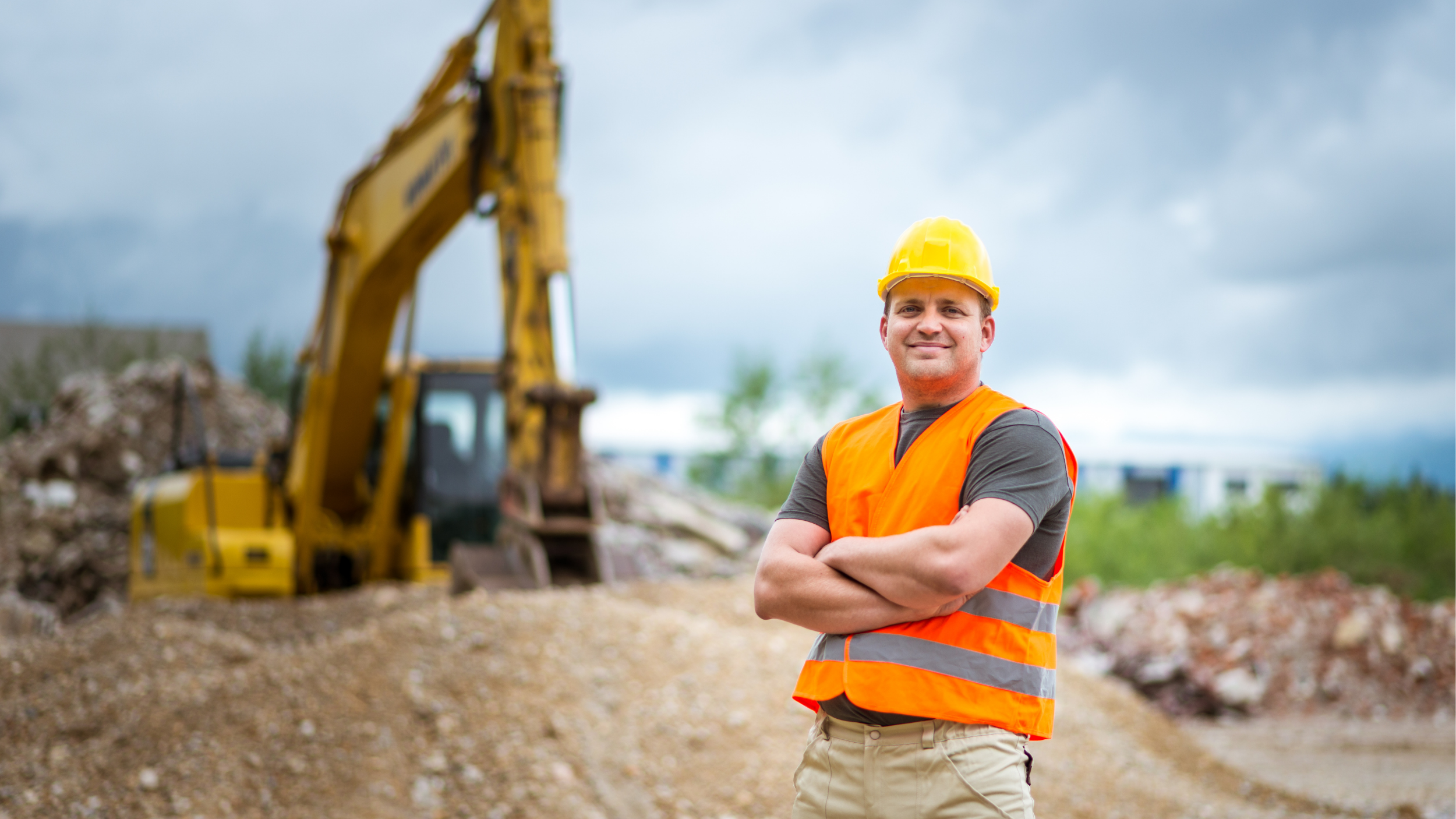 Construction worker wearing a yellow hard hat and orange safety vest stands with arms crossed at a job site, with an excavator and piles of dirt blurred in the background under a cloudy sky.