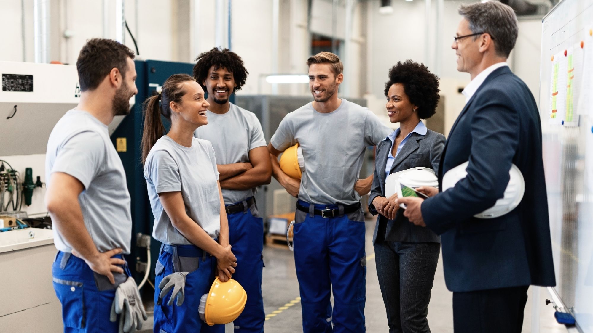 A group of industrial workers and supervisors stand together in a manufacturing facility during a team meeting, smiling and holding safety helmets while discussing operations near equipment and a planning board.