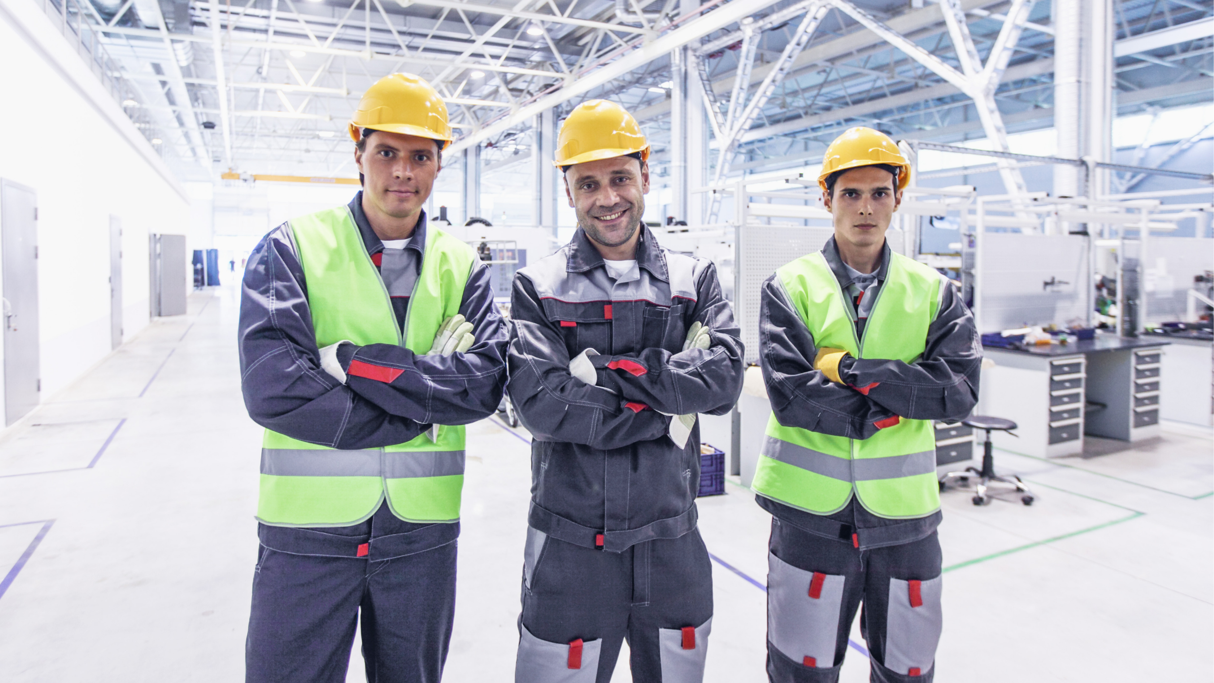 Three industrial workers wearing yellow hard hats, safety vests, and work uniforms stand with arms crossed inside a bright manufacturing facility.