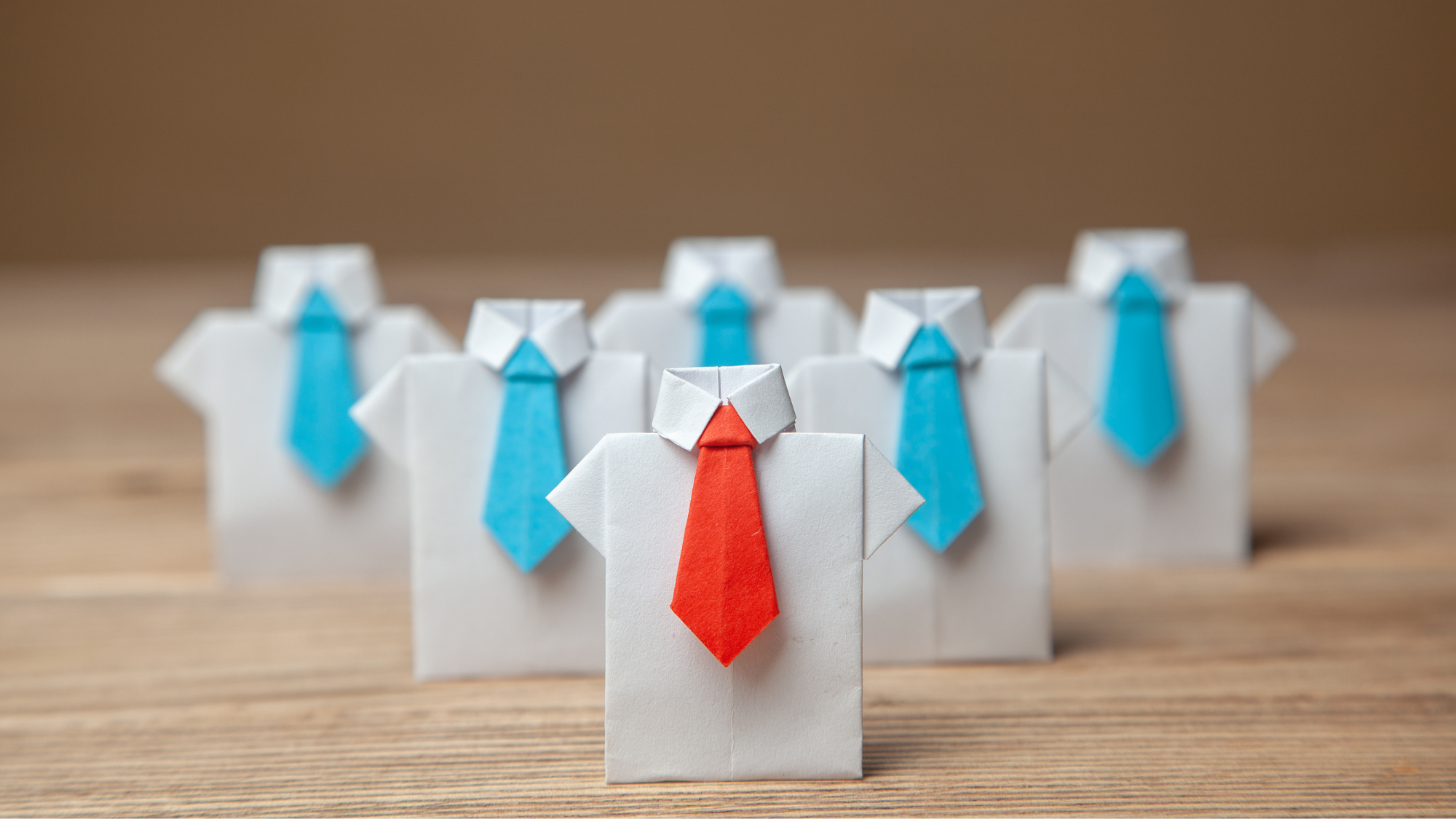 Paper origami figures shaped like business shirts and ties are arranged on a wooden surface, with one figure wearing a red tie standing in front of others wearing blue ties to symbolize leadership or a standout candidate.