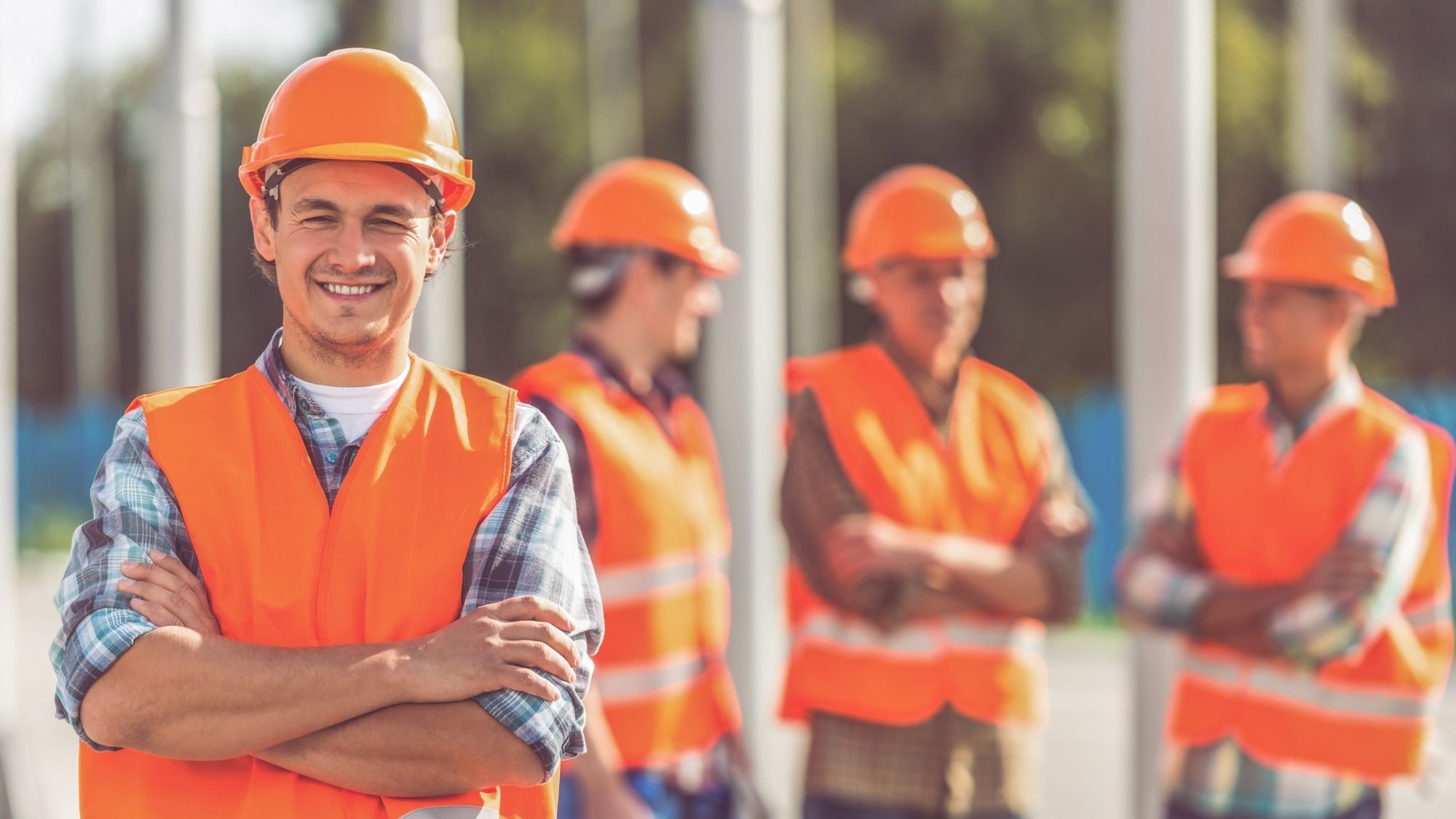 Smiling construction worker wearing an orange hard hat and reflective safety vest stands with arms crossed in the foreground, while three other workers in matching safety gear stand blurred in the background at a job site.