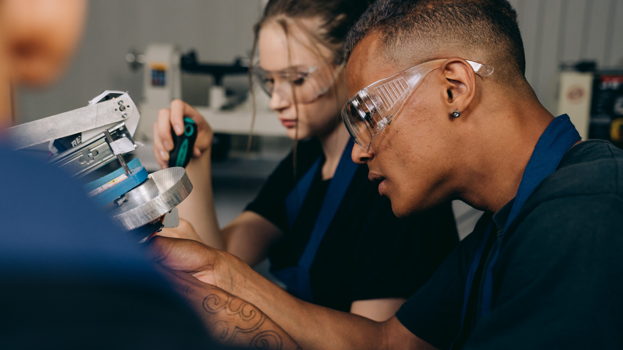 Two apprentices wearing safety glasses work together on a technical project in a workshop or training lab. One person carefully adjusts a machine component while the other observes and assists with a hand tool.
