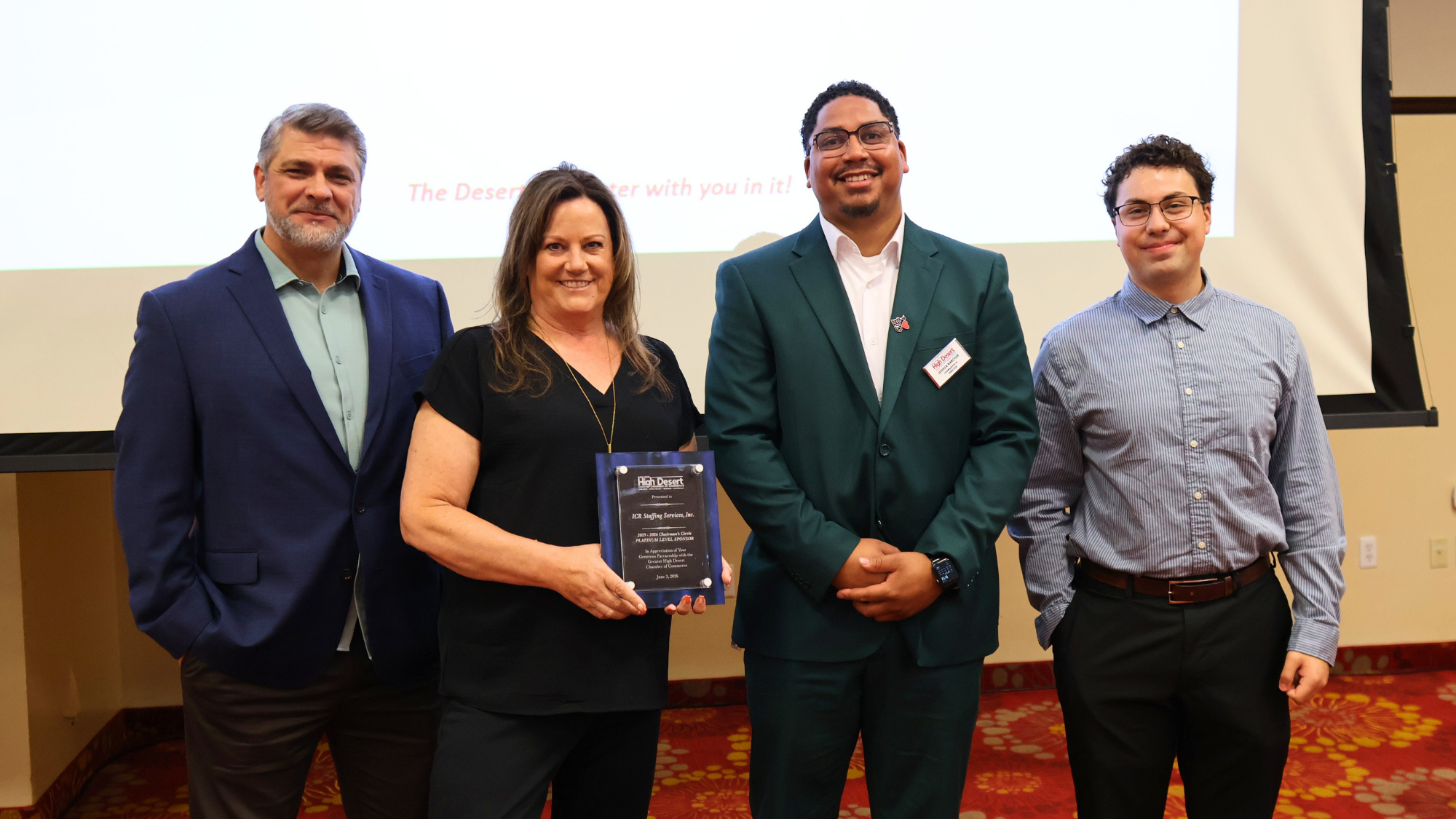 Four individuals pose together at a Greater High Desert Chamber of Commerce event. One woman holds a recognition plaque while standing between three colleagues. The group smiles for a photo in front of a presentation screen, celebrating an award or community achievement.