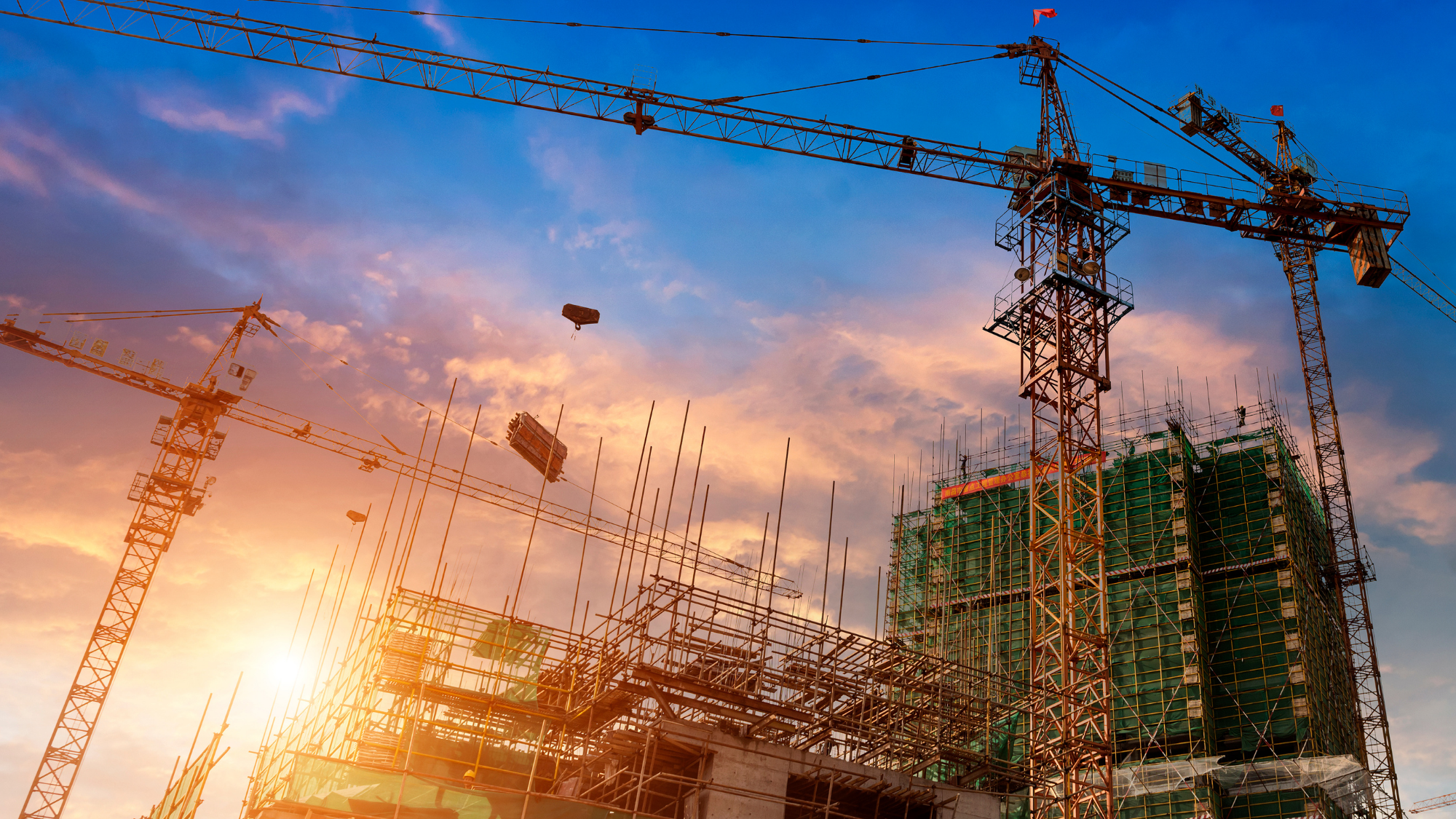 Large construction site at sunset featuring multiple tower cranes, steel framework, and scaffolding surrounding a multi-story building under development.