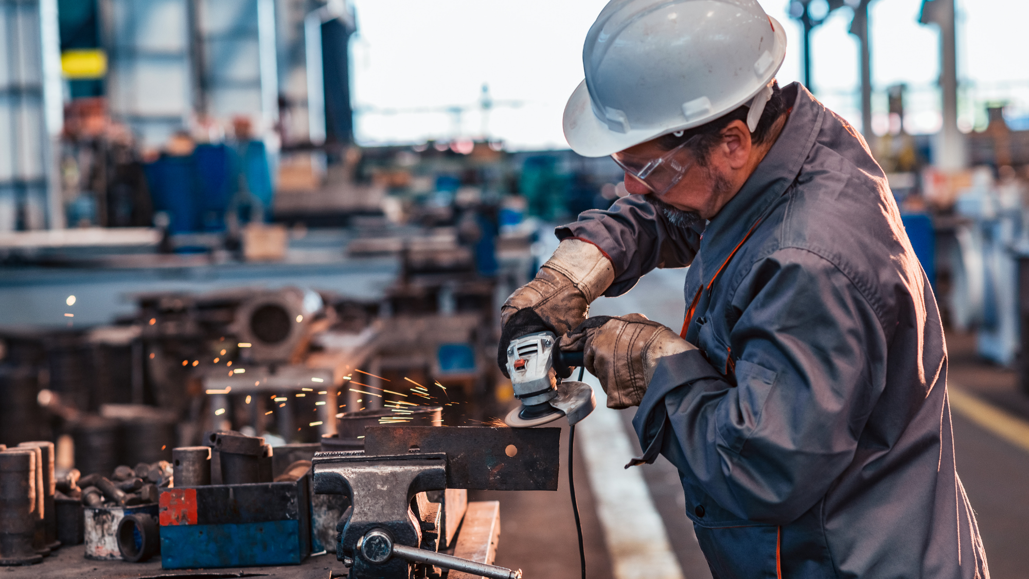 Industrial worker wearing a hard hat, safety glasses, gloves, and protective workwear uses an angle grinder on a metal component inside a manufacturing facility.