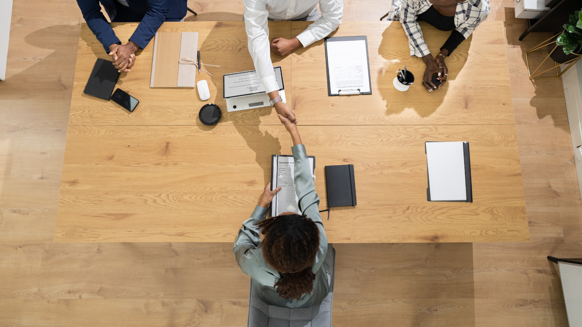 Overhead view of a job interview or hiring meeting where a candidate shakes hands with a hiring manager across a conference table while other interviewers observe, with resumes, folders, and office materials spread across the table.
