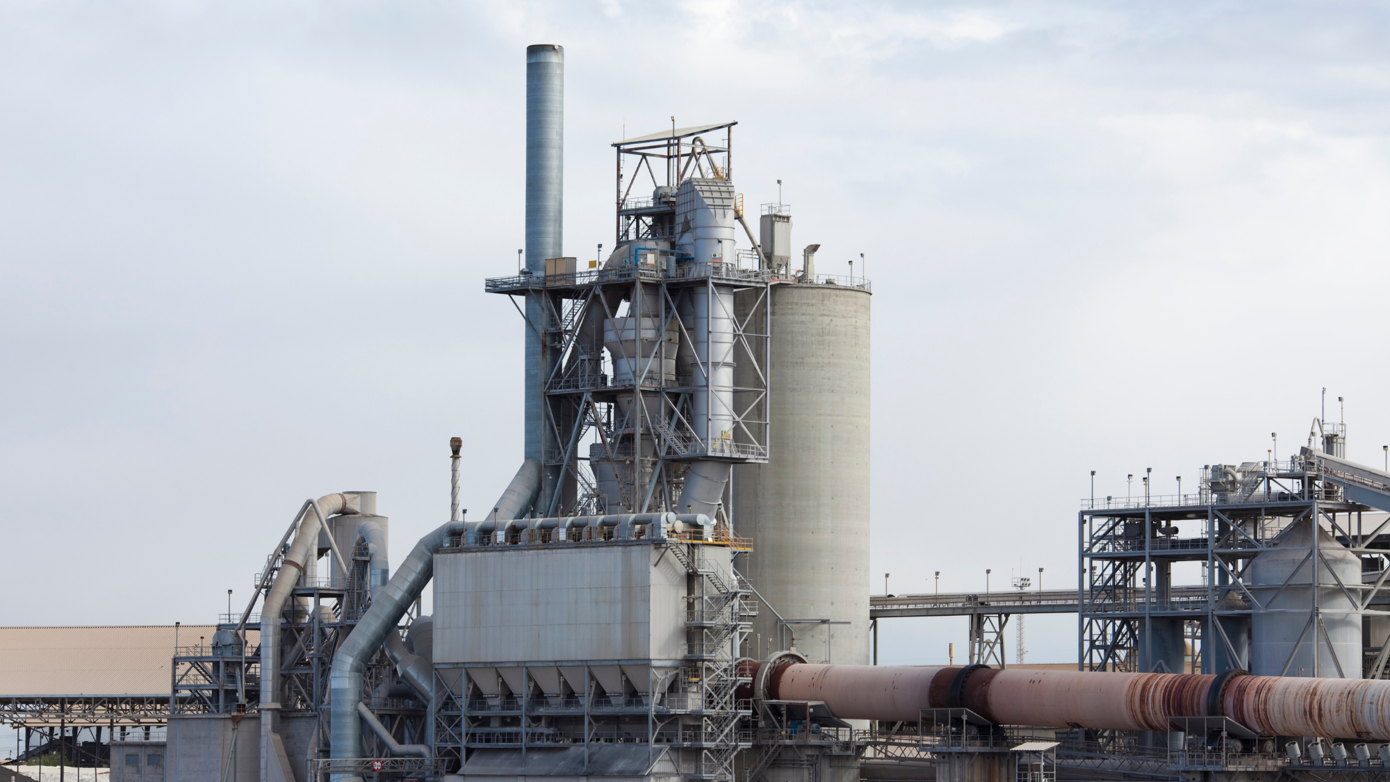 Industrial cement manufacturing facility featuring a large rotary kiln, cyclone preheater tower, smokestack, silos, conveyors, and interconnected processing equipment.