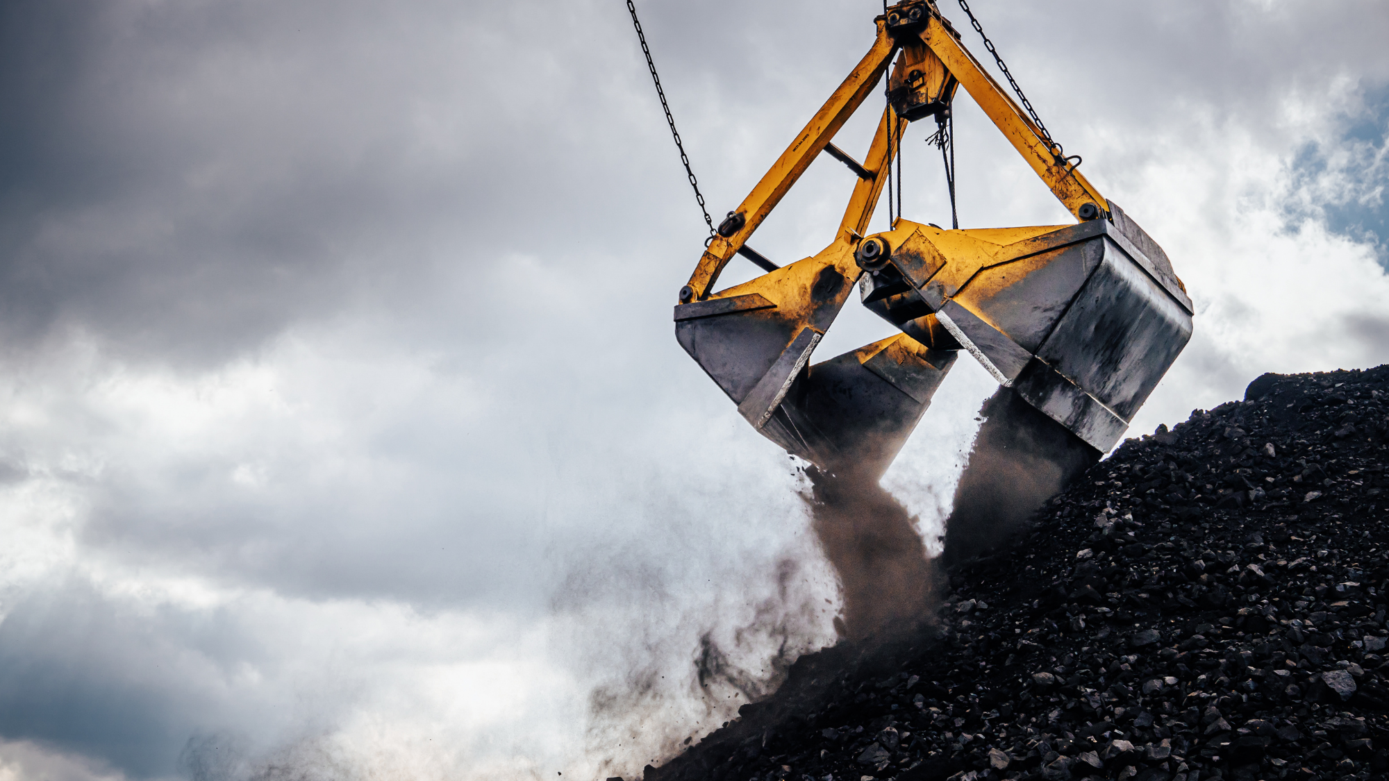 A large crane-mounted grab bucket releases a load of coal onto a stockpile at an industrial material handling facility.