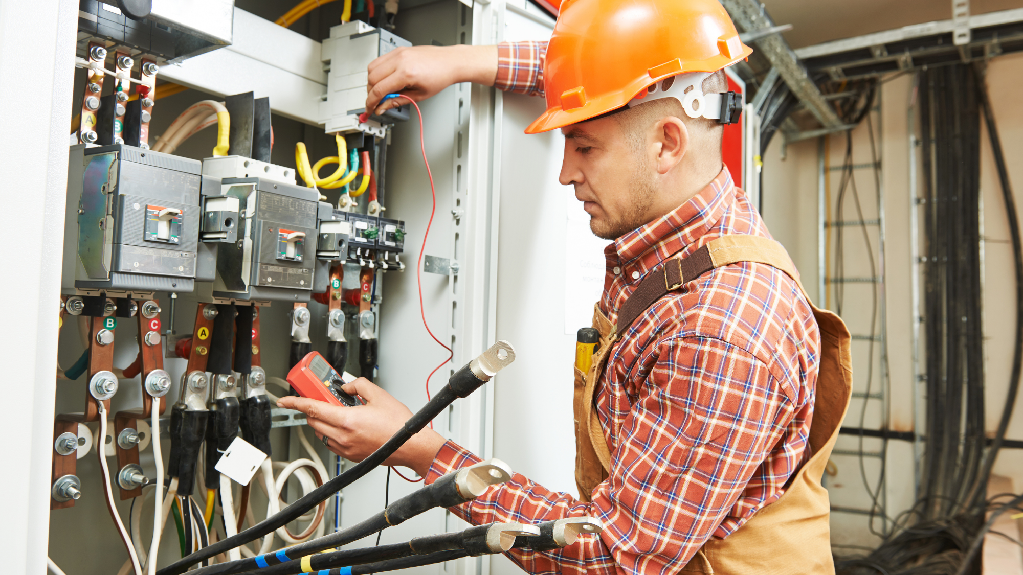 An electrician wearing an orange hard hat and safety gear tests an electrical control panel with a digital multimeter inside an industrial facility.
