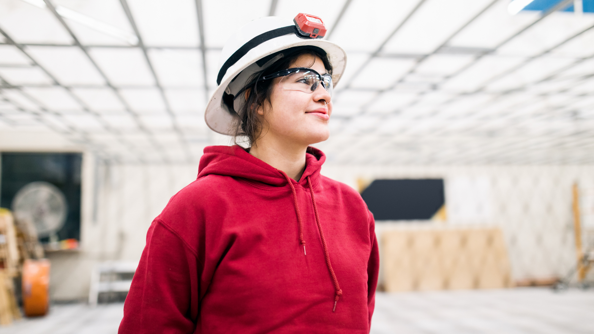 A student wearing a white hard hat with a headlamp, clear safety glasses, and a red hoodie stands inside an industrial facility, looking confidently into the distance.