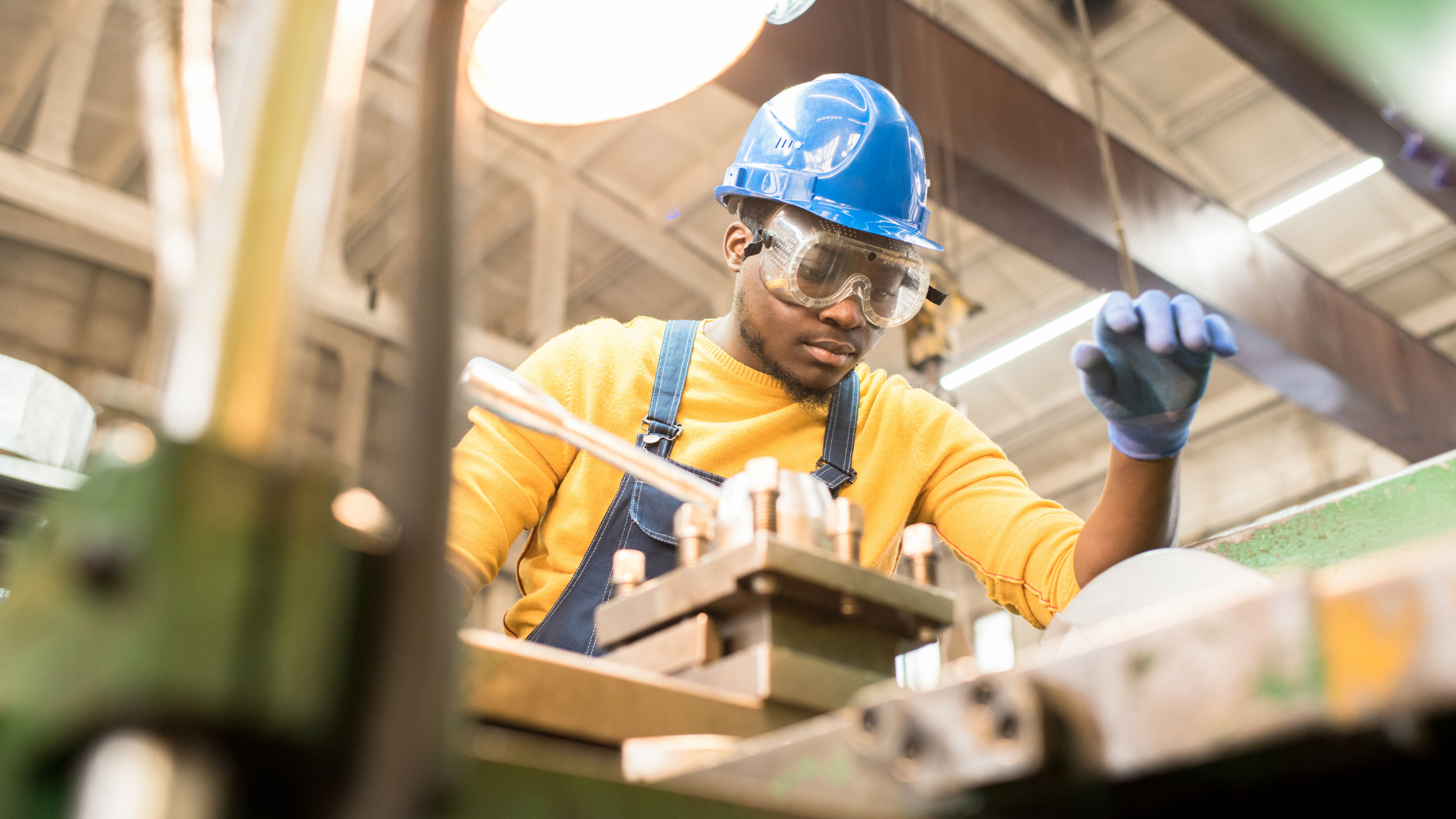 Worker wearing a blue hard hat, safety goggles, gloves, and overalls operating industrial machinery inside a manufacturing facility.
