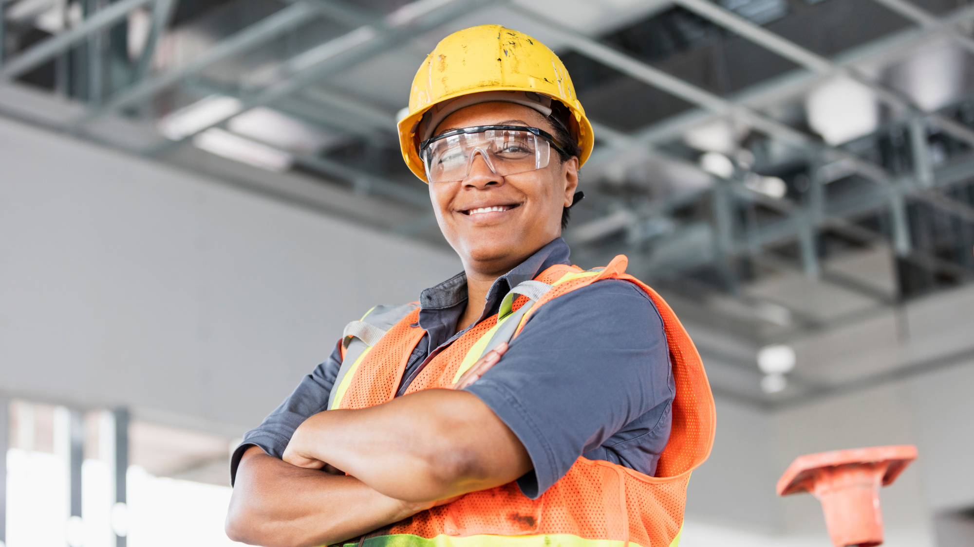 Smiling construction worker wearing a yellow hard hat, safety glasses, and a high-visibility safety vest stands with arms crossed inside a commercial building under construction.