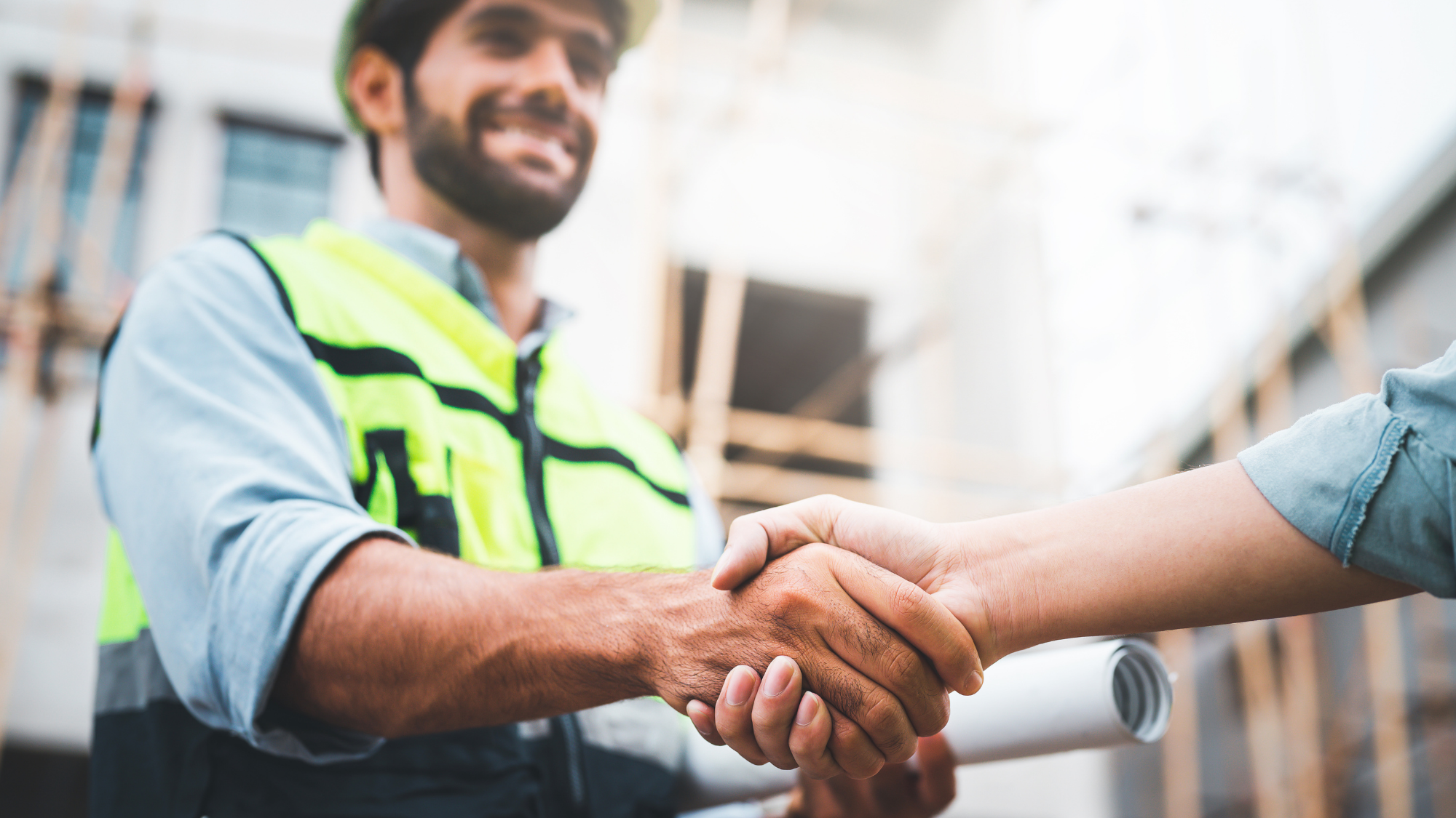 Close-up of a handshake with a smiling industrial worker wearing a hard hat and high-visibility safety vest in the background.