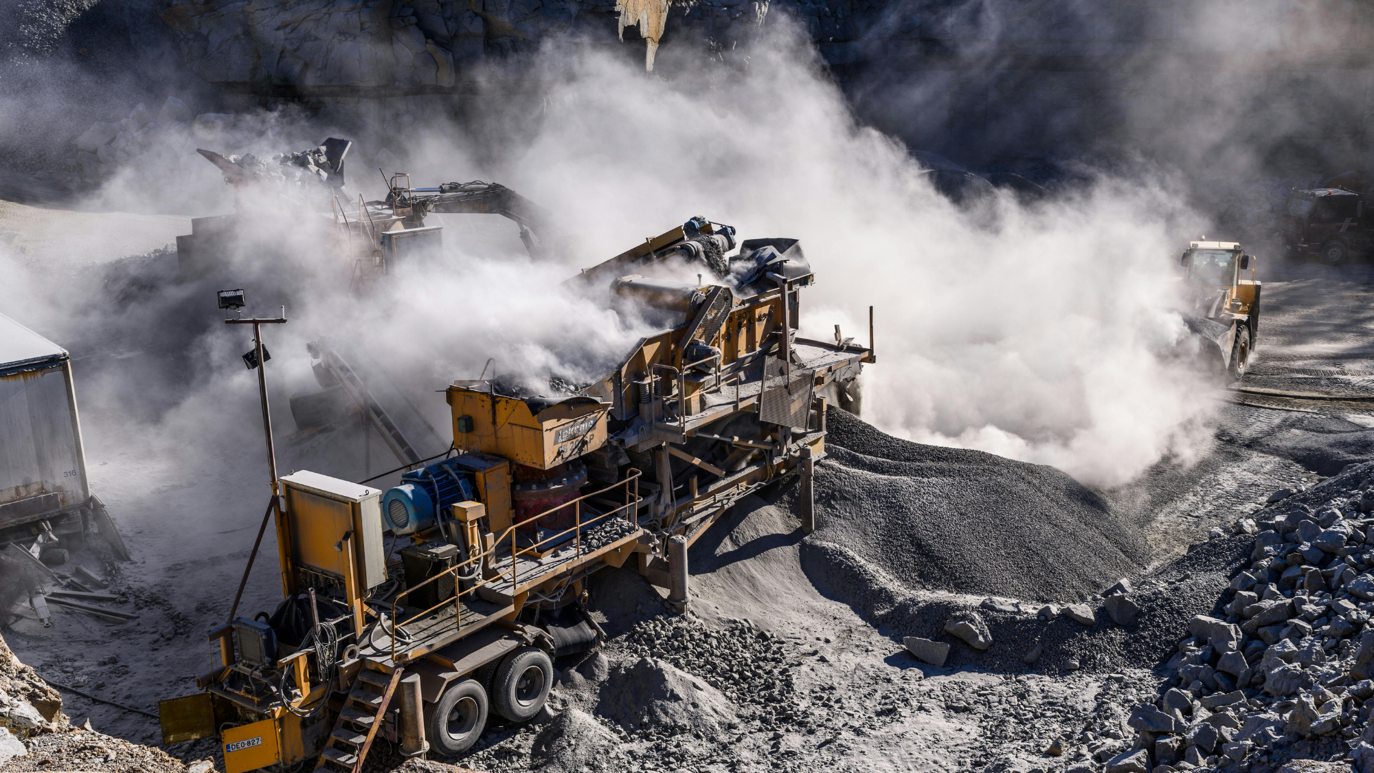 Portable rock crushing equipment processes aggregate at an active quarry, creating large clouds of dust as crushed stone is discharged onto stockpiles.