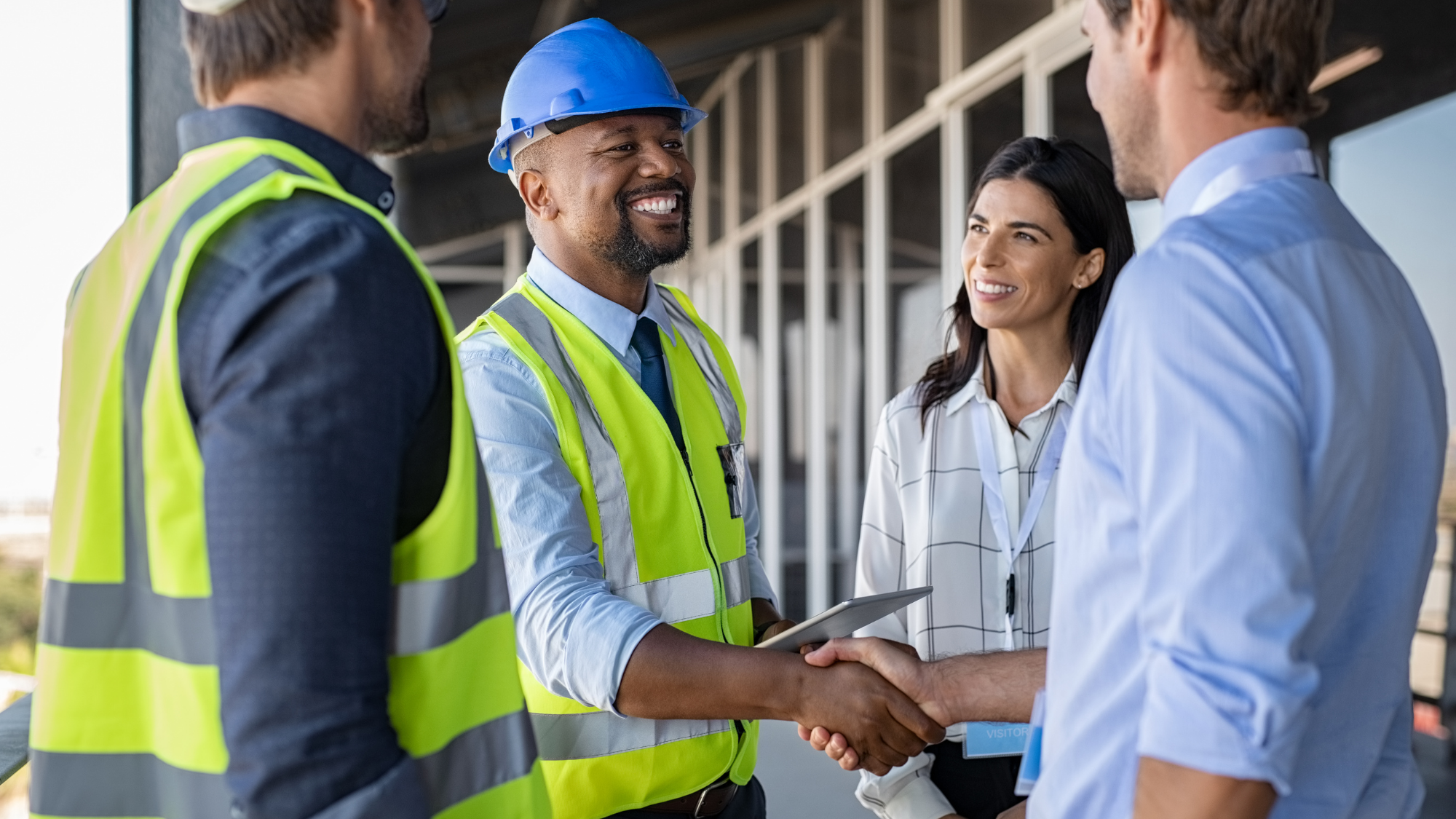 Industrial professionals wearing high-visibility safety vests and hard hats greet business representatives with a handshake at a construction site.