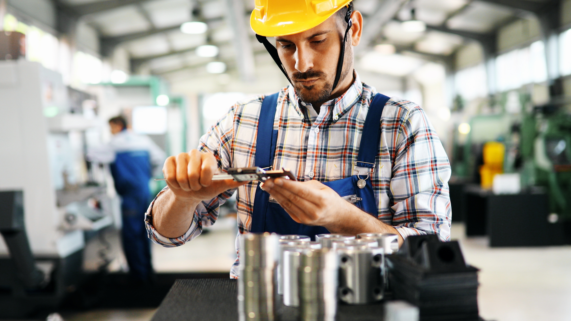 Skilled manufacturing technician wearing a hard hat and safety gear uses a caliper to measure a precision-machined metal component in a modern industrial workshop.