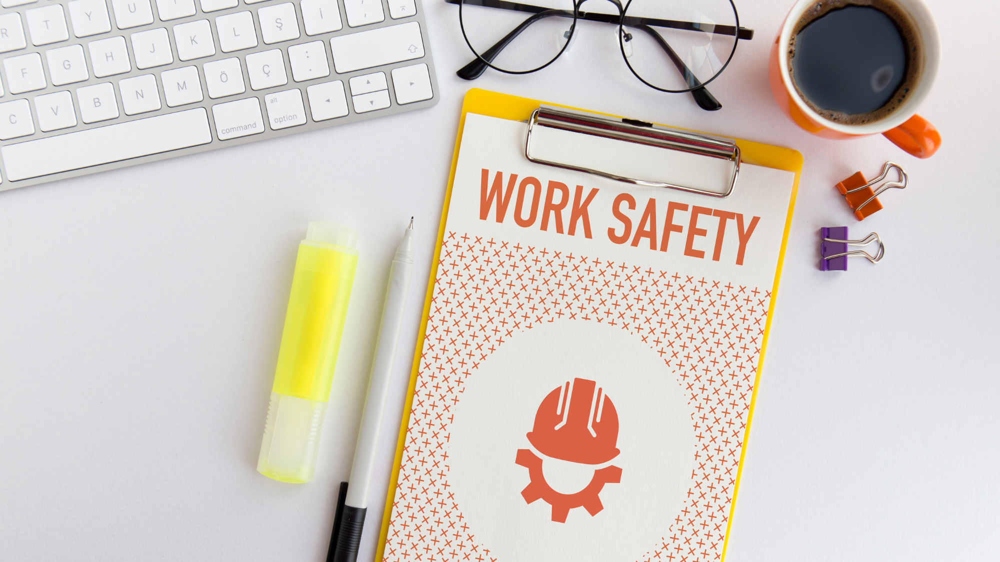 A clipboard with a piece of paper that reads "Work Safety," featuring an icon of a construction worker. A cup of coffee, a keyboard, glasses, and a highlighter are next to the clipboard.