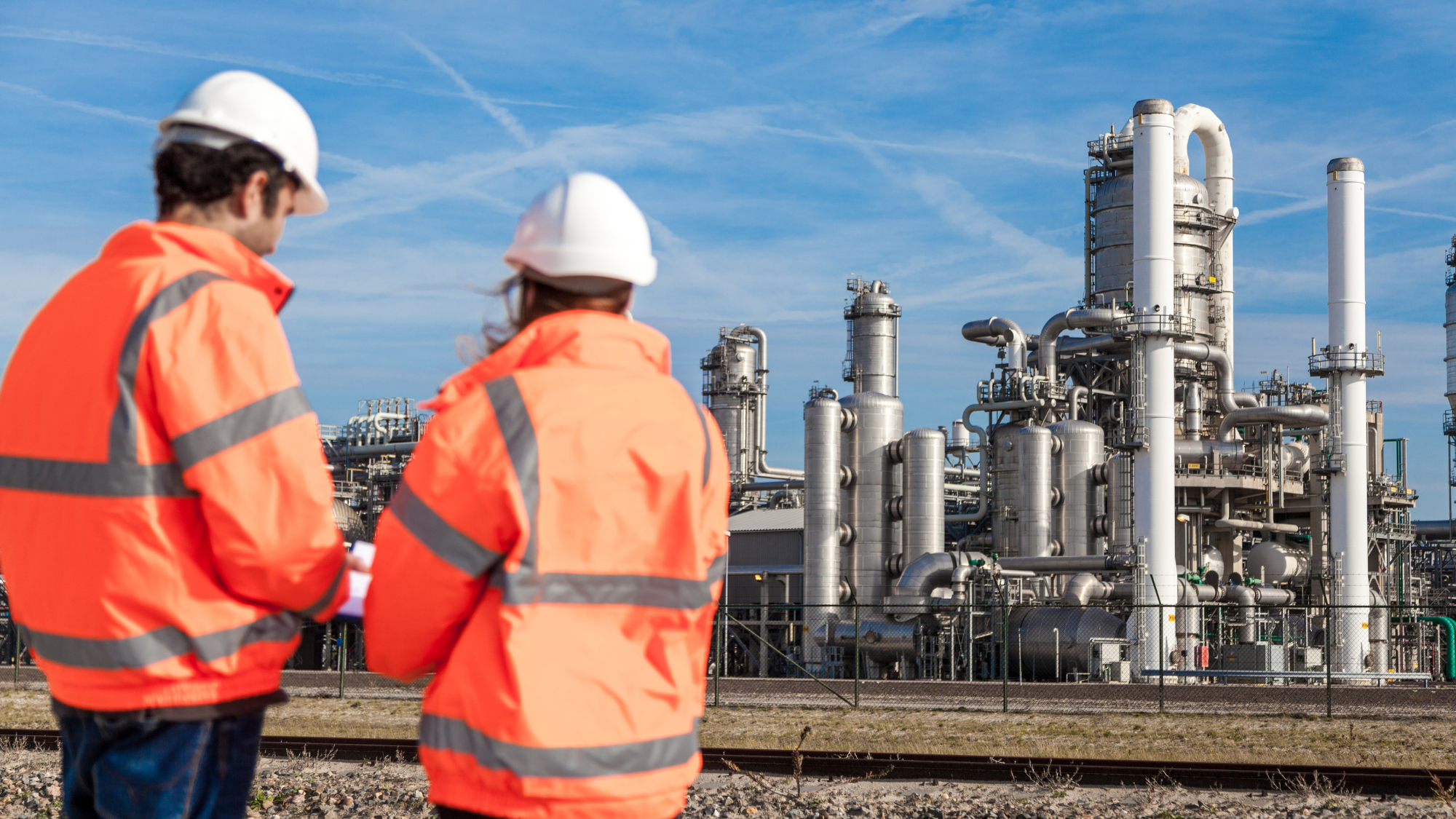Two industrial workers wearing white hard hats and orange high-visibility jackets look over a large processing facility filled with towers, pipes, tanks, and other heavy industrial equipment.