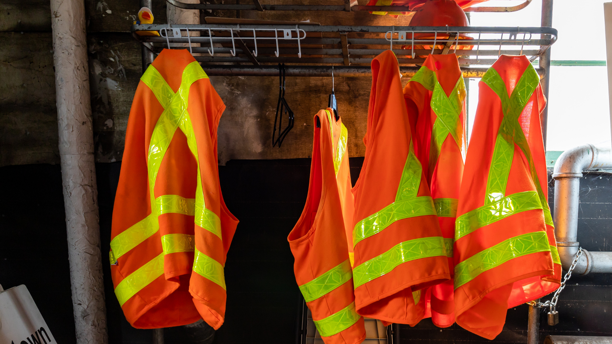 Bright orange high-visibility safety vests with reflective yellow stripes hang on a rack inside an industrial work area, ready for workers to use.