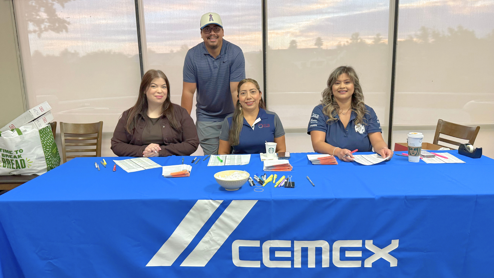 Four people smile behind a CEMEX-branded registration table with paperwork, pens, and event materials at a company or community event.