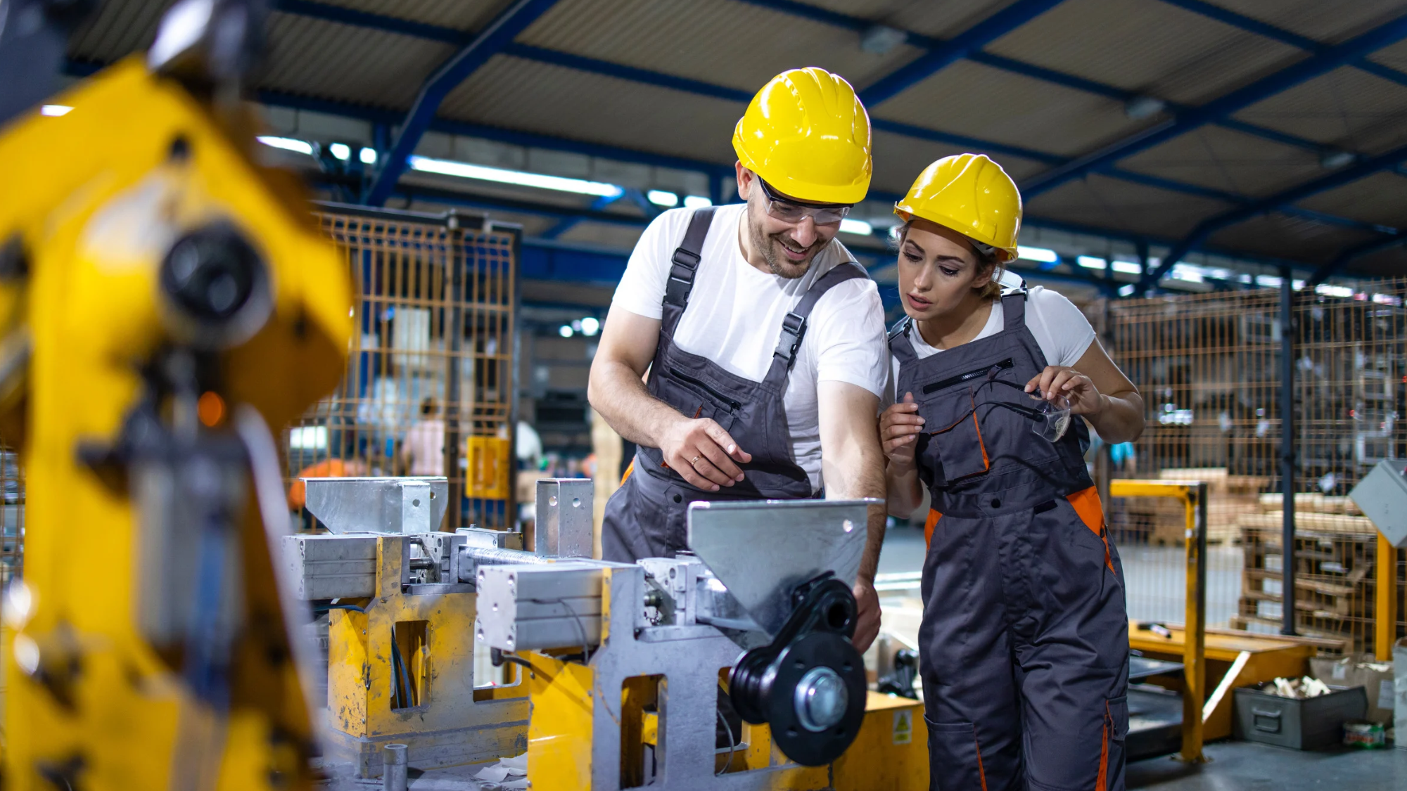 Two industrial workers wearing hard hats and safety gear inspect machinery together on a manufacturing floor.