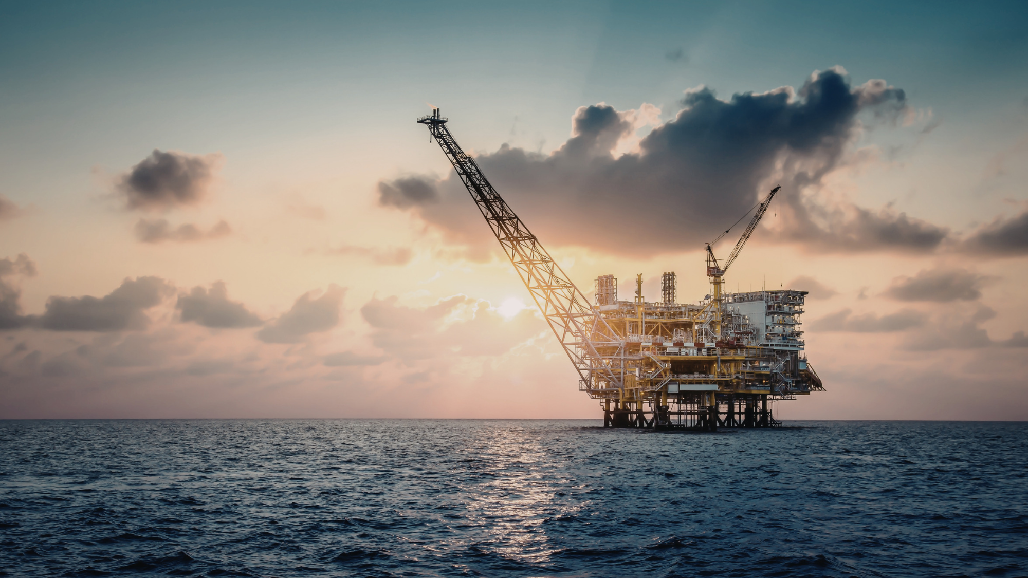 Offshore mining platform with large cranes standing in the ocean at sunset beneath a dramatic cloudy sky.